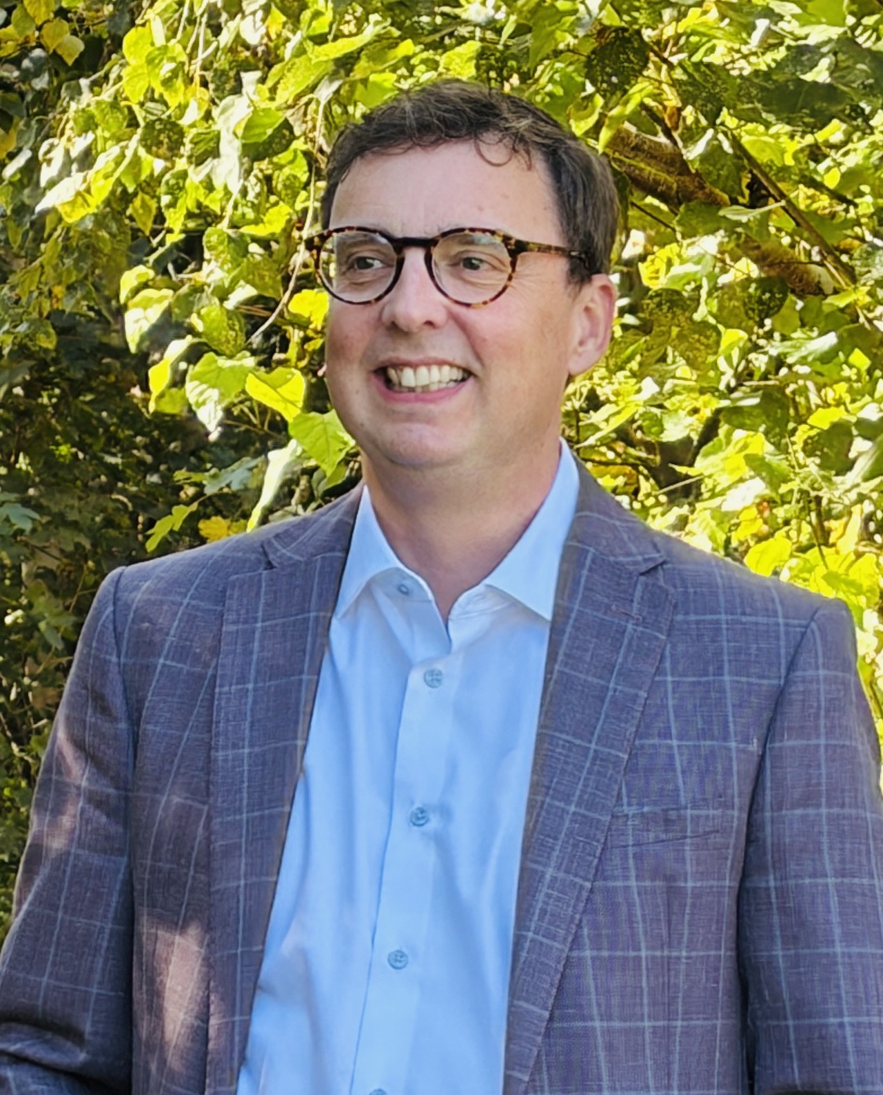 Scott Barfoot, smiling, in a grey checked blazer and light blue shirt outdoors in front of green leaves